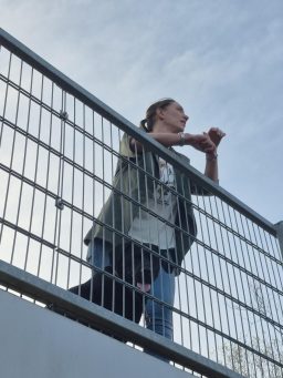 Eine Person steht an einem Balkon und spricht, mit einer offenen Handbewegung.