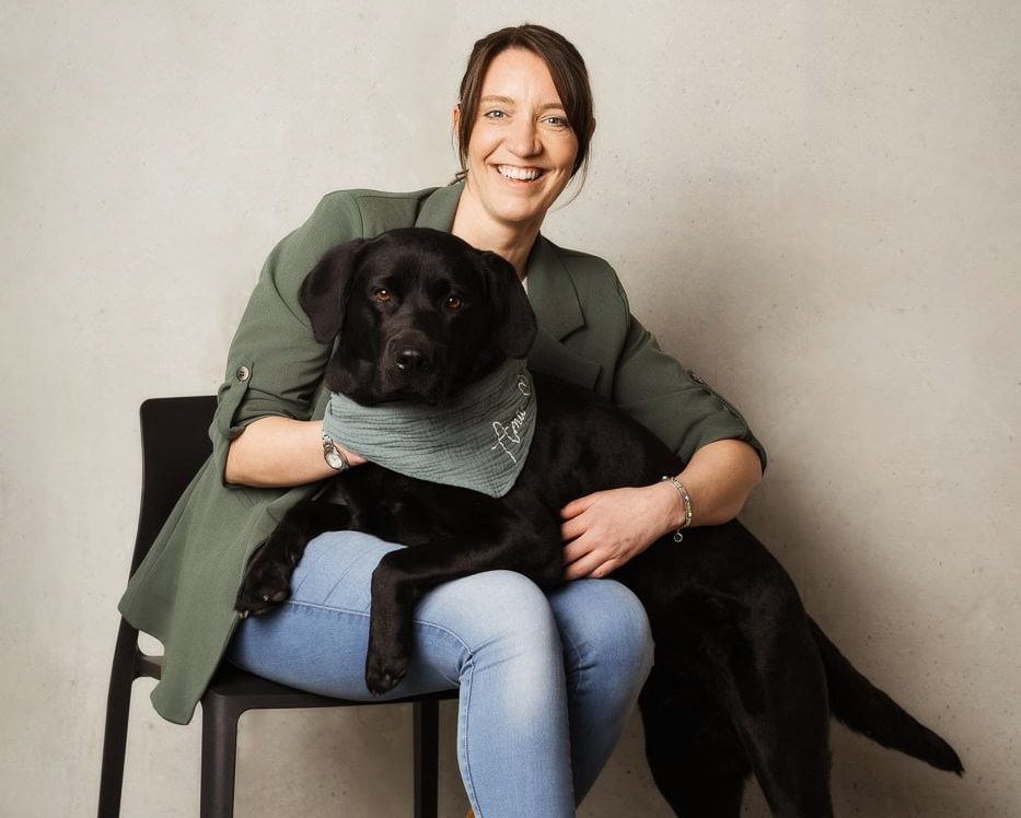 Maria Röming mit ihrer schwarzen Labrador (Amie) sitzt auf einem Stuhl und lächelt.