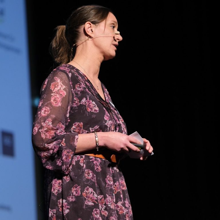 Frau in floral Kleid spricht auf einer Bühne, Publikum im Hintergrund.